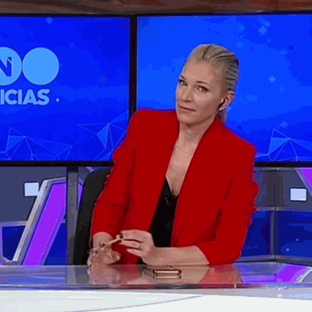 a woman in a red jacket sits at a desk in front of a screen that says " noticias "