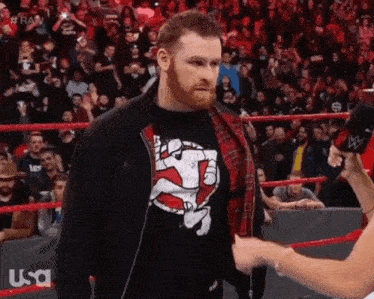 a man in a black shirt with a ghostbusters logo on it stands in a wrestling ring