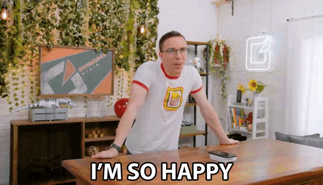a man wearing a taco bell shirt is standing in front of a table and says i 'm so happy