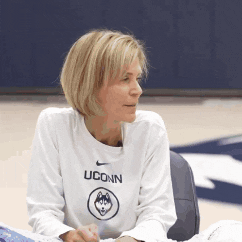 a woman wearing a white uconn shirt sits on a chair