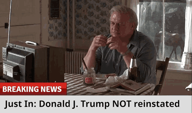 a man sits at a table in front of a television with a breaking news banner above him