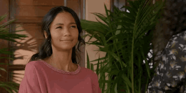 a woman in a pink shirt is smiling while standing next to a plant .