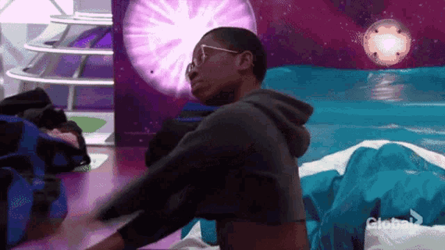 a man in a hoodie and glasses is standing in front of a purple wall with the word global on it .
