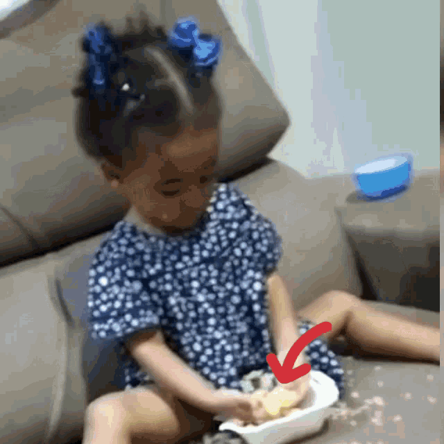 a little girl is sitting on a couch playing with a bowl of food with an arrow pointing to it