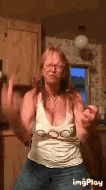 a woman is dancing in a kitchen wearing a white tank top and jeans .