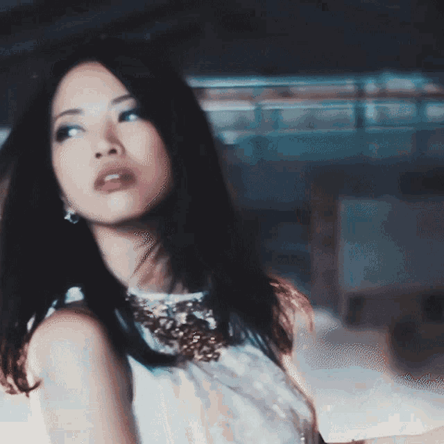 a woman with long black hair is wearing a white top with a necklace around her neck