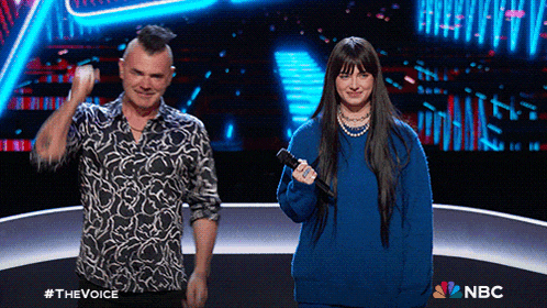a man with a mohawk and a woman in a blue sweater stand on a stage in front of a nbc sign