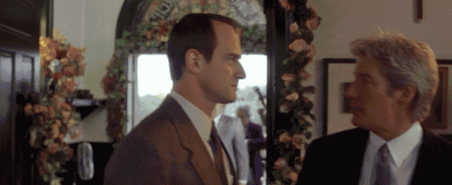 two men in suits and ties are talking to each other in a room with flowers in the background .