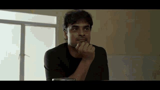 a man in a black shirt is sitting in front of a window with his hand on his chin