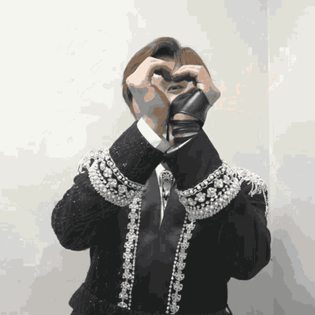 a man wearing a black jacket with pearls on the sleeves making a heart shape with his hands