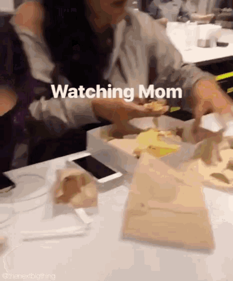 a woman is sitting at a table with a box of food and a cell phone while watching mom
