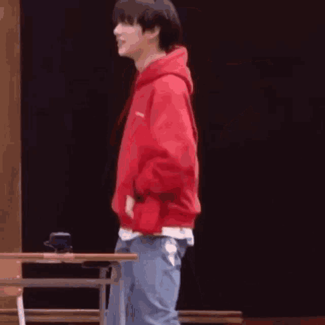 a man in a red hoodie and jeans is standing in front of a desk .