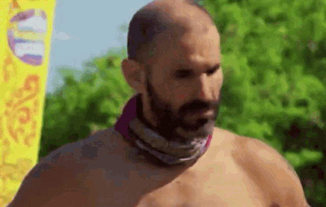 a shirtless man with a beard and a scarf around his neck is standing in front of a yellow banner .