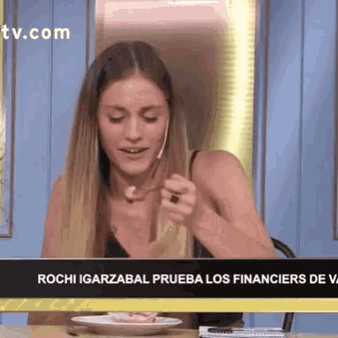 a woman is sitting at a table with a plate of food in front of a tv screen that says rochi garzabal