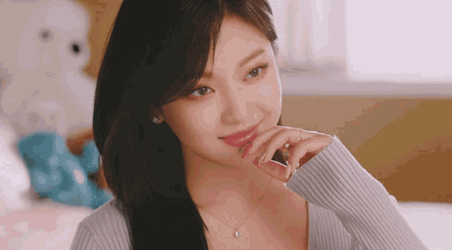 a woman wearing a sweater and a necklace smiles with her hand on her chin