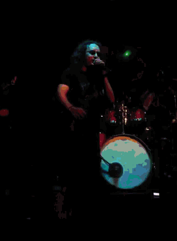 a man singing into a microphone in front of a drum set in a dark room