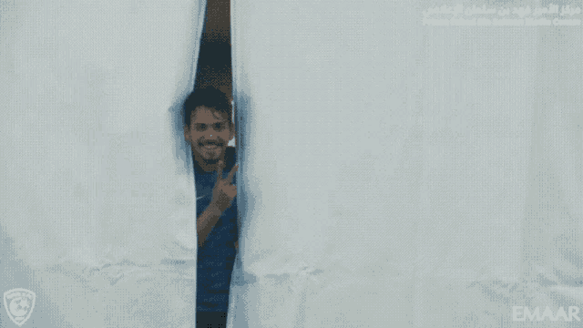 a man in a blue shirt is peeking out from behind a white curtain with emaar written on the bottom