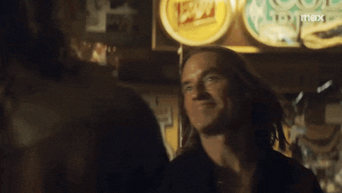 a man with long hair is smiling in front of a sign that says ' beer ' on it .