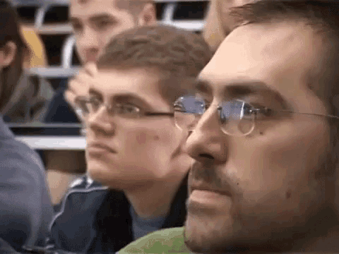 a man wearing glasses is sitting in a lecture hall with other people .