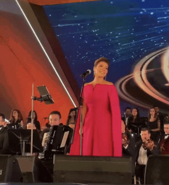 a woman in a pink dress sings into a microphone in front of an orchestra