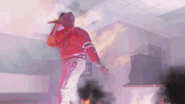 a man in an orange jersey is singing into a microphone in front of a fire