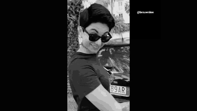a black and white photo of a young man wearing glasses and a choker standing next to a car .