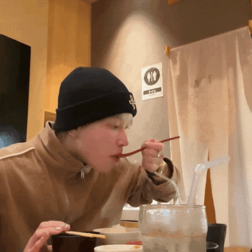 a man wearing a black beanie is eating with chopsticks in front of a sign that says toilet