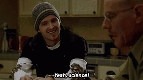 two men are sitting at a table in a kitchen talking to each other . one of the men is wearing a beanie .