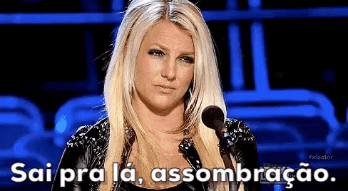 a blonde woman is standing in front of a microphone with the words sai pra la assombracao written above her .