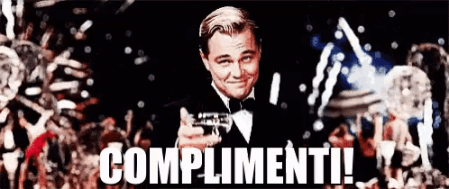 a man in a tuxedo is holding a bottle of champagne and smiling with the words complimenti written below him .