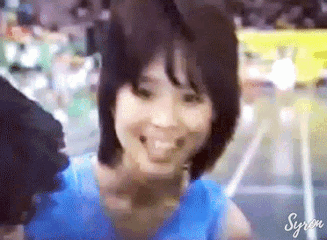 a woman in a blue shirt is smiling in a blurry photo with syron written below her