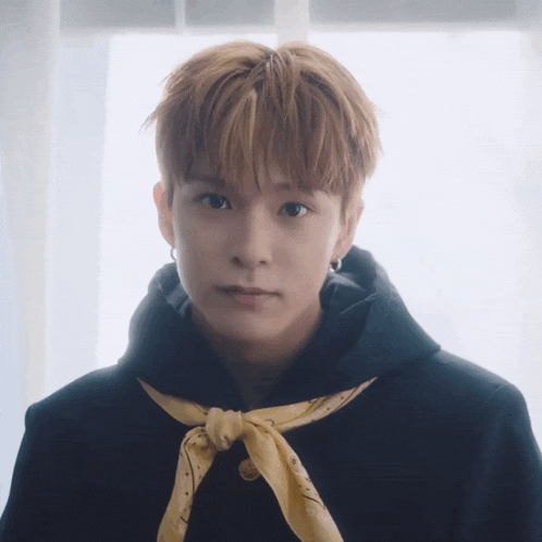 a young man wearing a black hoodie and a yellow scarf around his neck looks at the camera
