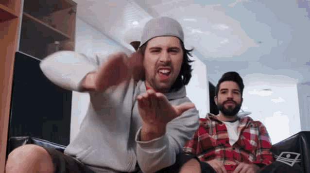 a man with a beard is sitting on a couch making a funny face while another man watches .