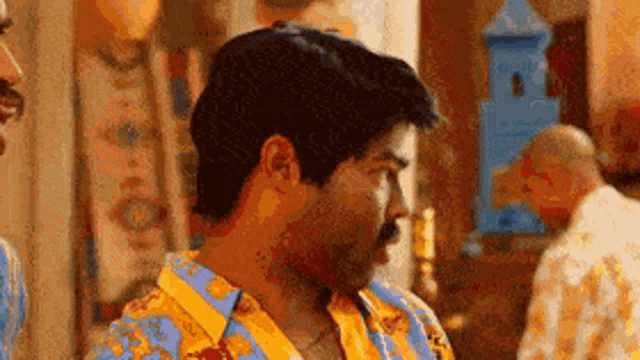 a man with a mustache is wearing a colorful shirt and talking to another man in a room .