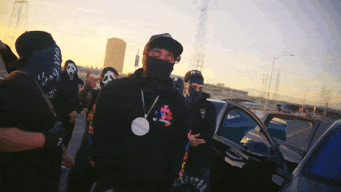 a man wearing a black hoodie with a smiley face on it stands in front of a car