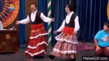 two men in long skirts are dancing on a stage in front of a wheel of fortune .