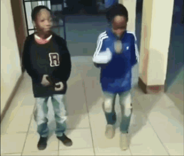 two young boys are standing next to each other in a hallway . one of the boys has the letter r on his shirt .