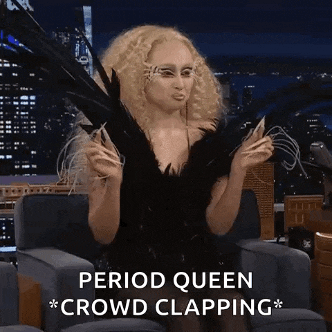 a woman in a black dress with feathers on her face is sitting in a chair with the words period queen crowd clapping .