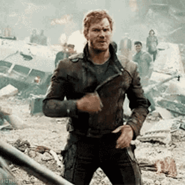 a man in a leather jacket is standing in a destroyed city holding a gun .