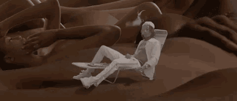 a man in a white jacket is sitting in a chair in front of a pile of naked women .