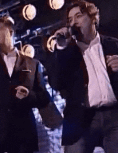 a man in a suit sings into a microphone while another man looks on