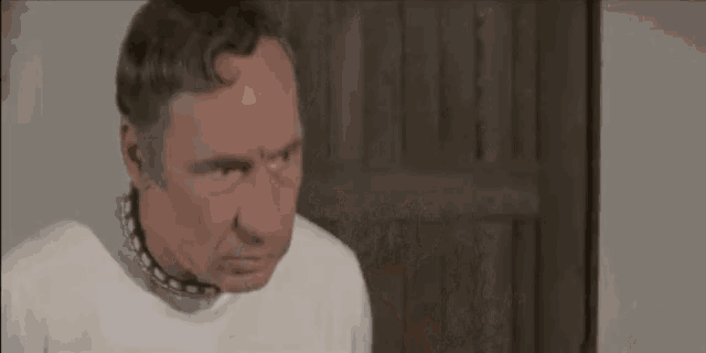 a man wearing a white shirt and a chain around his neck is standing in front of a wooden door .