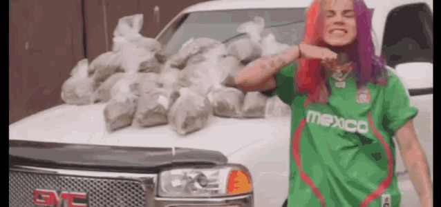 a woman with purple hair and a green shirt is standing in front of a gmc truck filled with bags of drugs .