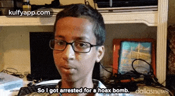 a young boy wearing glasses is sitting in front of a computer desk and talking about being arrested for a hoax bomb .