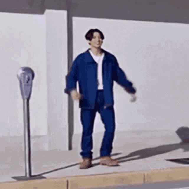 a man in a blue jacket and jeans is standing on the sidewalk next to a parking meter .