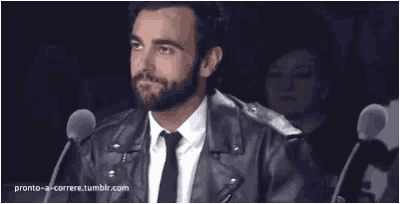 a man with a beard wearing a leather jacket and tie is sitting in front of microphones ..