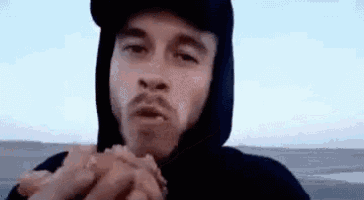 a man in a hooded jacket is eating a hamburger on the beach .