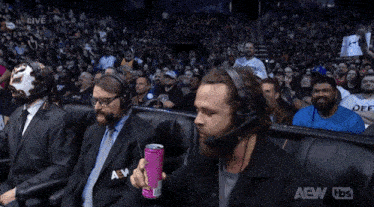a group of men are sitting in a stadium watching a wrestling match sponsored by aew tbs .