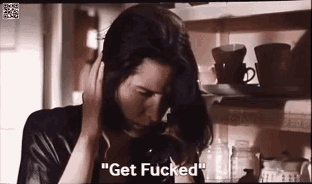 a woman is talking on a cell phone in a kitchen while holding her head and saying `` get fucked '' .
