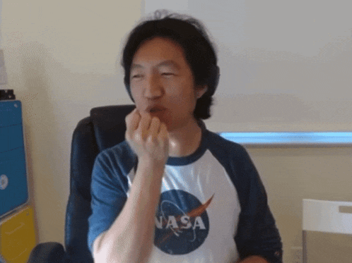 a man wearing a nasa shirt sits in a chair with his hand in his mouth